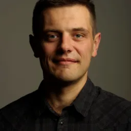 Portret mężczyzny z krótkimi włosami w ciemnej koszuli w kratę, uśmiechającego się delikatnie do kamery na szarym tle.