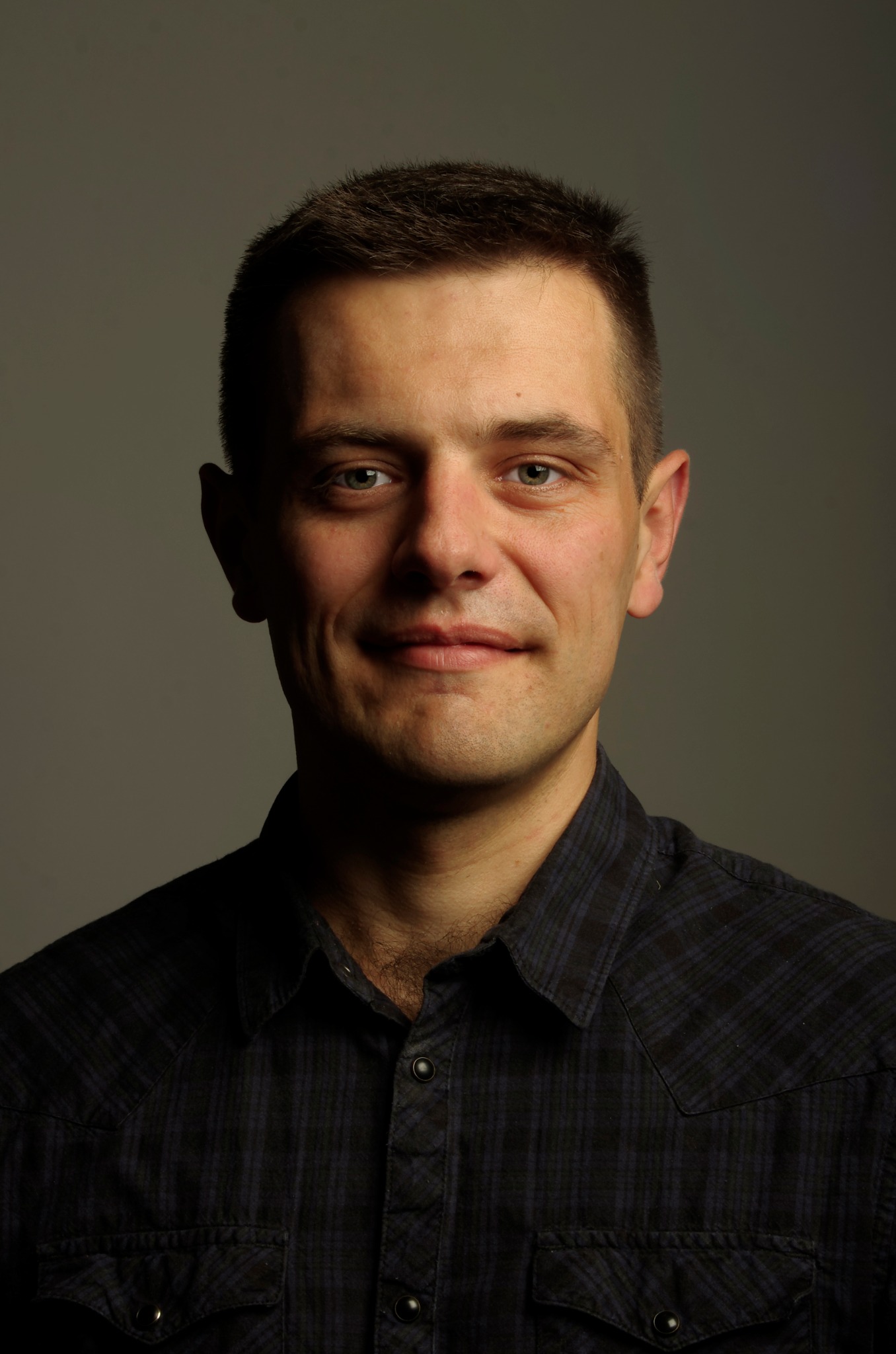 Portret mężczyzny z krótkimi włosami w ciemnej koszuli w kratę, uśmiechającego się delikatnie do kamery na szarym tle.