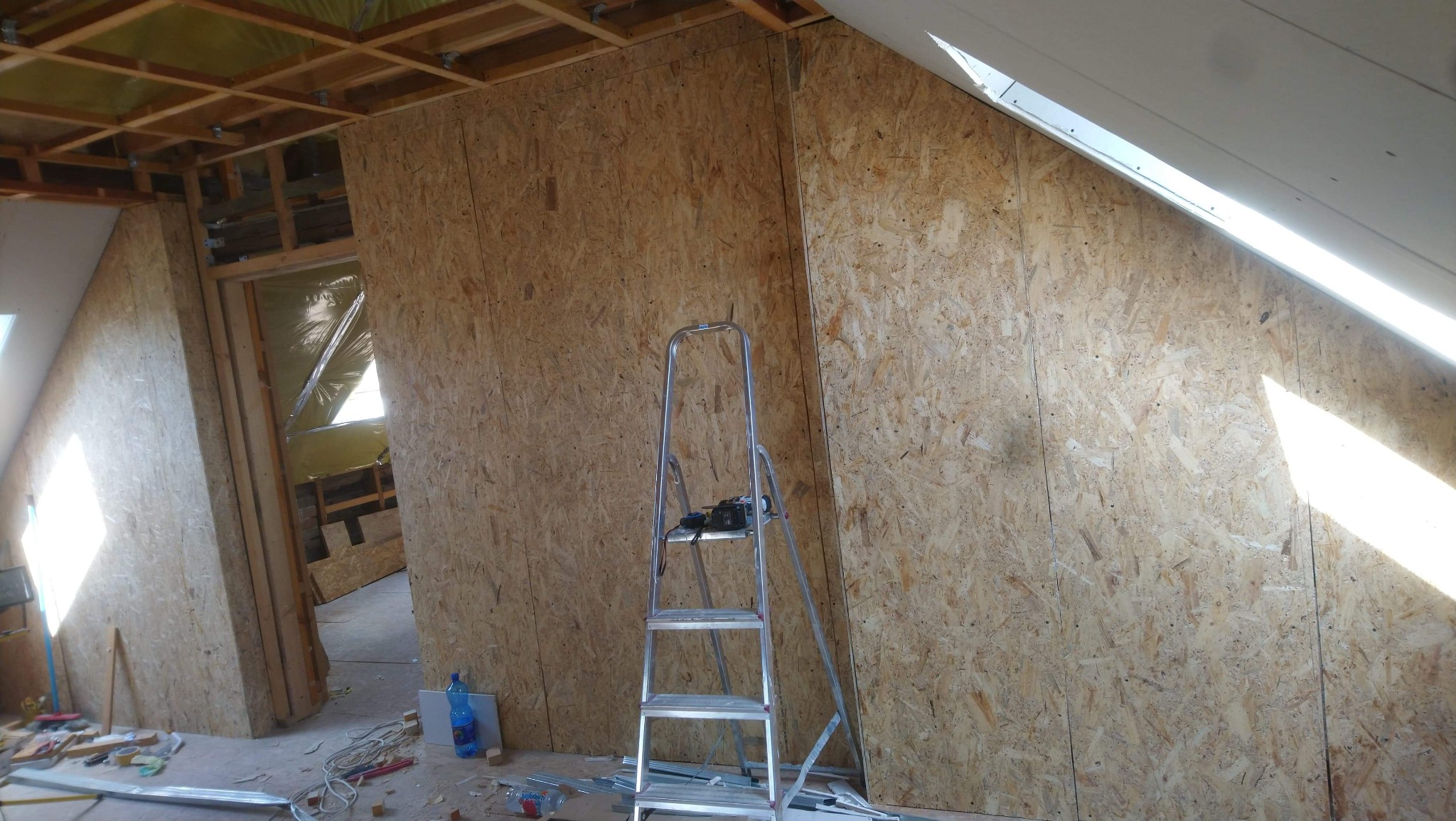 Montaż ścian z płyty OSB na poddaszu, widoczna konstrukcja drewniana dachu, izolacja, drabina aluminiowa i rozłożone narzędzia.