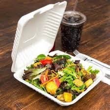 Sałatka w białym, ekologicznym pudełku na wynos, obok napój w plastikowym kubku z rurką i sztućce na drewnianym stole.