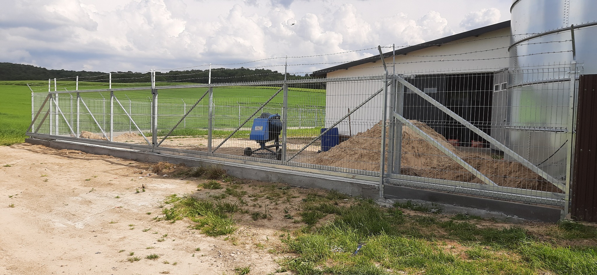 Przesuwna brama z siatki, zabezpieczona drutem kolczastym, widoczna na tle zielonego pola i budynku gospodarczego, z betoniarką i stertą piasku wewnątrz ogrodzenia.