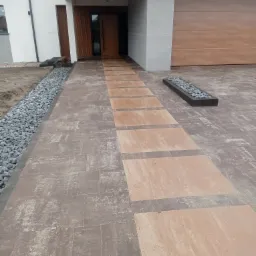 Nowoczesna posesja z wybrukowaną ścieżką z prostokątnych płyt w dwóch odcieniach brązu, prowadzącą do drewnianych drzwi wejściowych, z dekoracyjnym pasem kamieni po lewej stronie i minimalistyczną...