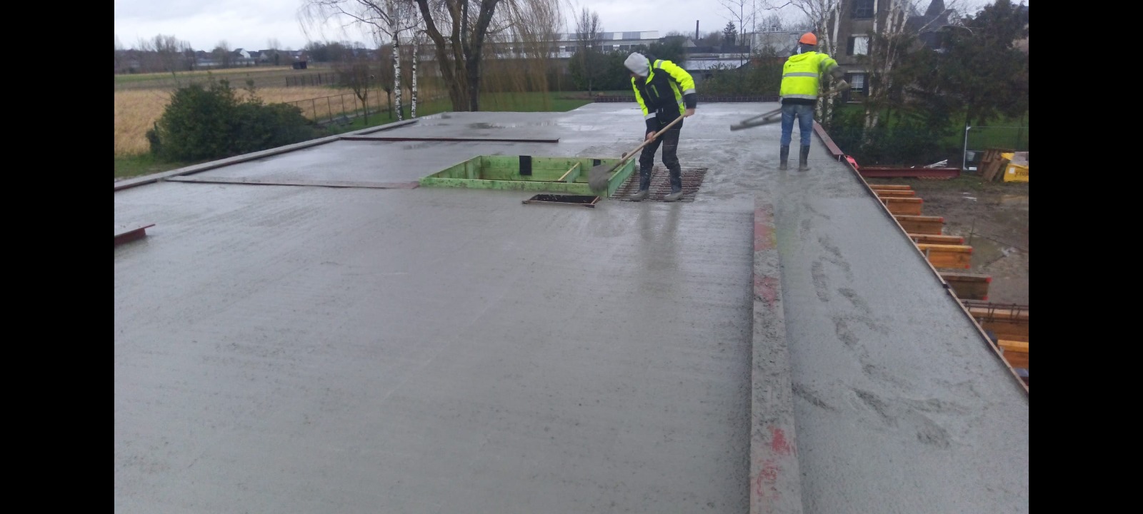 Wylewka betonowa na stropie budynku w trakcie prac wykończeniowych; dwóch robotników w odblaskowych ubraniach rozprowadza beton za pomocą narzędzi, widoczne szalunki na obrzeżach stropu, elementy...