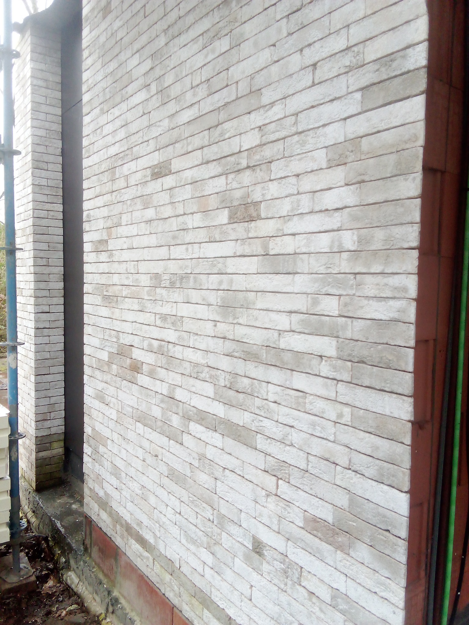 Ściana budynku wykończona jasnoszarą cegłą klinkierową, widoczny fragment rusztowania i narożnik budynku z brązową ramą okienną.