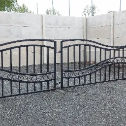 Czarna, metalowa brama dwuskrzydłowa z ozdobnymi elementami w kształcie spirali, ustawiona na żwirowym podłożu przed betonowym ogrodzeniem modułowym.