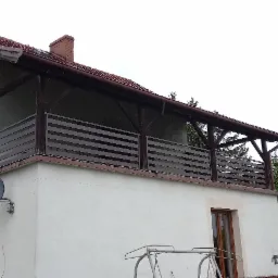 Dwupiętrowy dom z dużym balkonem z poziomą, brązową balustradą i drewnianą konstrukcją nośną dachu, widok z dołu w pochmurny dzień.