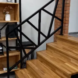 Nowoczesna balustrada schodowa z czarnej stali o geometrycznym wzorze, zamontowana przy drewnianych schodach z dębowymi stopniami, w tle regał z dekoracjami i fragment ściany z cegły.