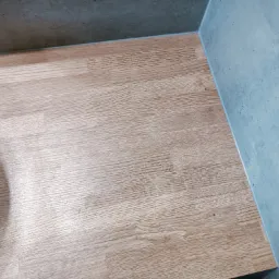 Fragment łazienki z jasną podłogą imitującą drewno i szarą ścianą z betonu architektonicznego, widoczny fragment białej ceramiki sanitarnej.