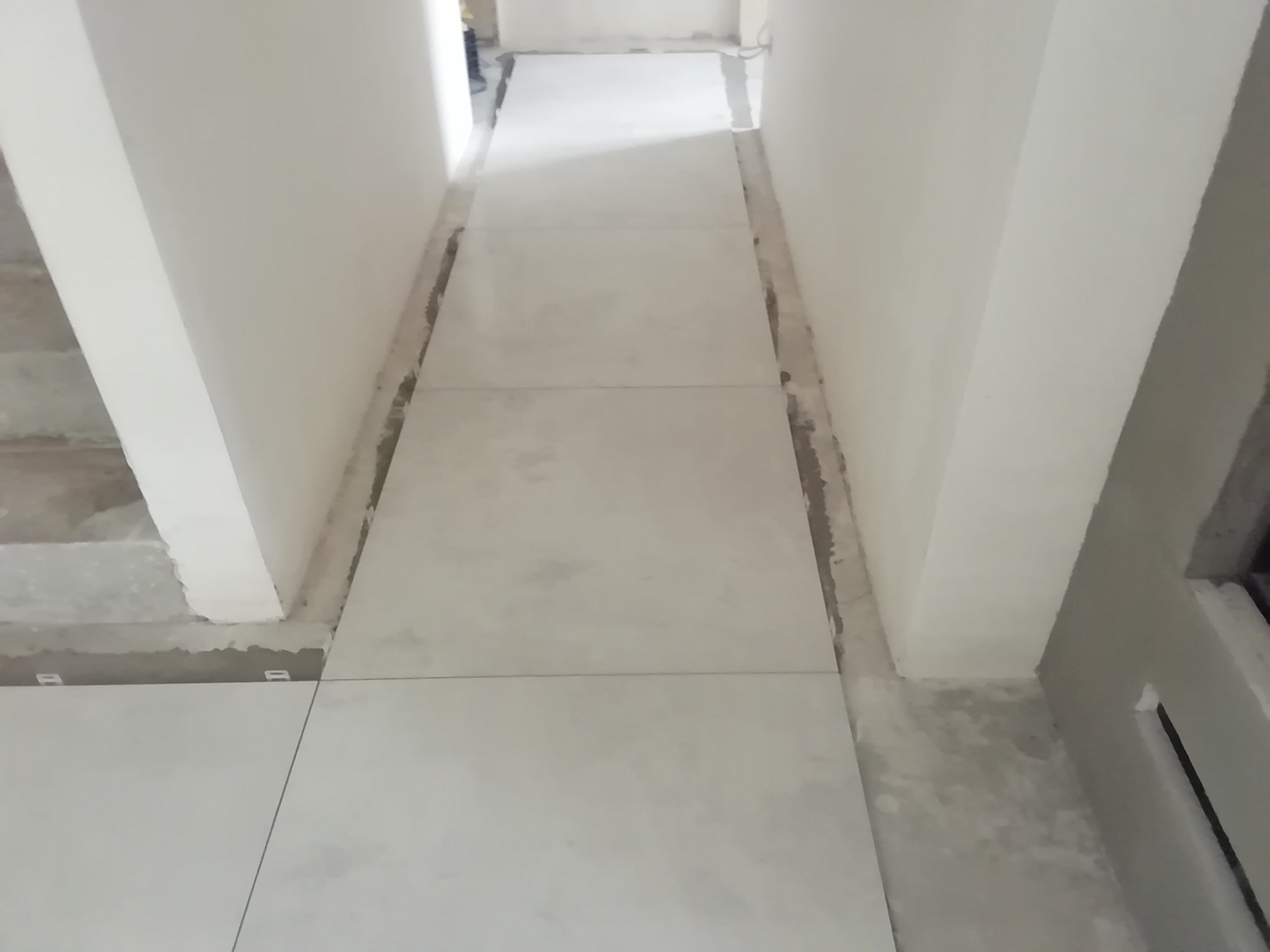 Układanie jasnych, kwadratowych płytek podłogowych w wąskim korytarzu, widoczne białe ściany i fragment schodów w tle, szare fugi pomiędzy płytkami.