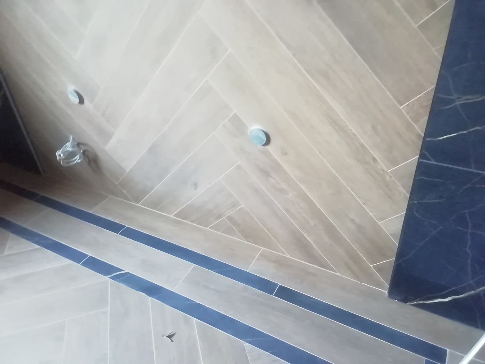 Ułożona w jodełkę beżowa podłoga z płytek w Warszawie, z ciemnoniebieskim pasem dekoracyjnym i fragmentem czarnego blatu imitującego marmur.