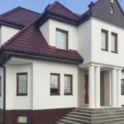 Elewacja domu jednorodzinnego po remoncie, z białymi ścianami, brązową dachówką i ceglanym cokołem, widoczne kolumny przy wejściu i schody.