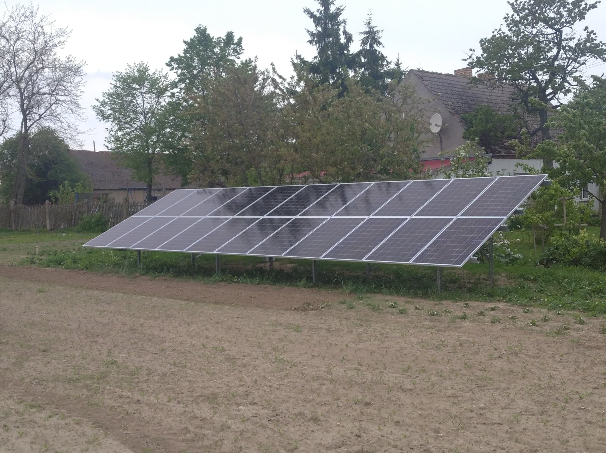 Instalacja fotowoltaiczna na gruncie w Koninie, składająca się z kilkunastu modułów, widoczna na tle wiejskiego krajobrazu z drzewami i budynkami.