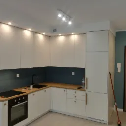 Nowoczesna kuchnia w odcieniach bieli i drewna z ciemnoniebieskim panelem nad blatem, oświetlona punktowymi lampami sufitowymi, z widocznymi narzędziami na blacie.