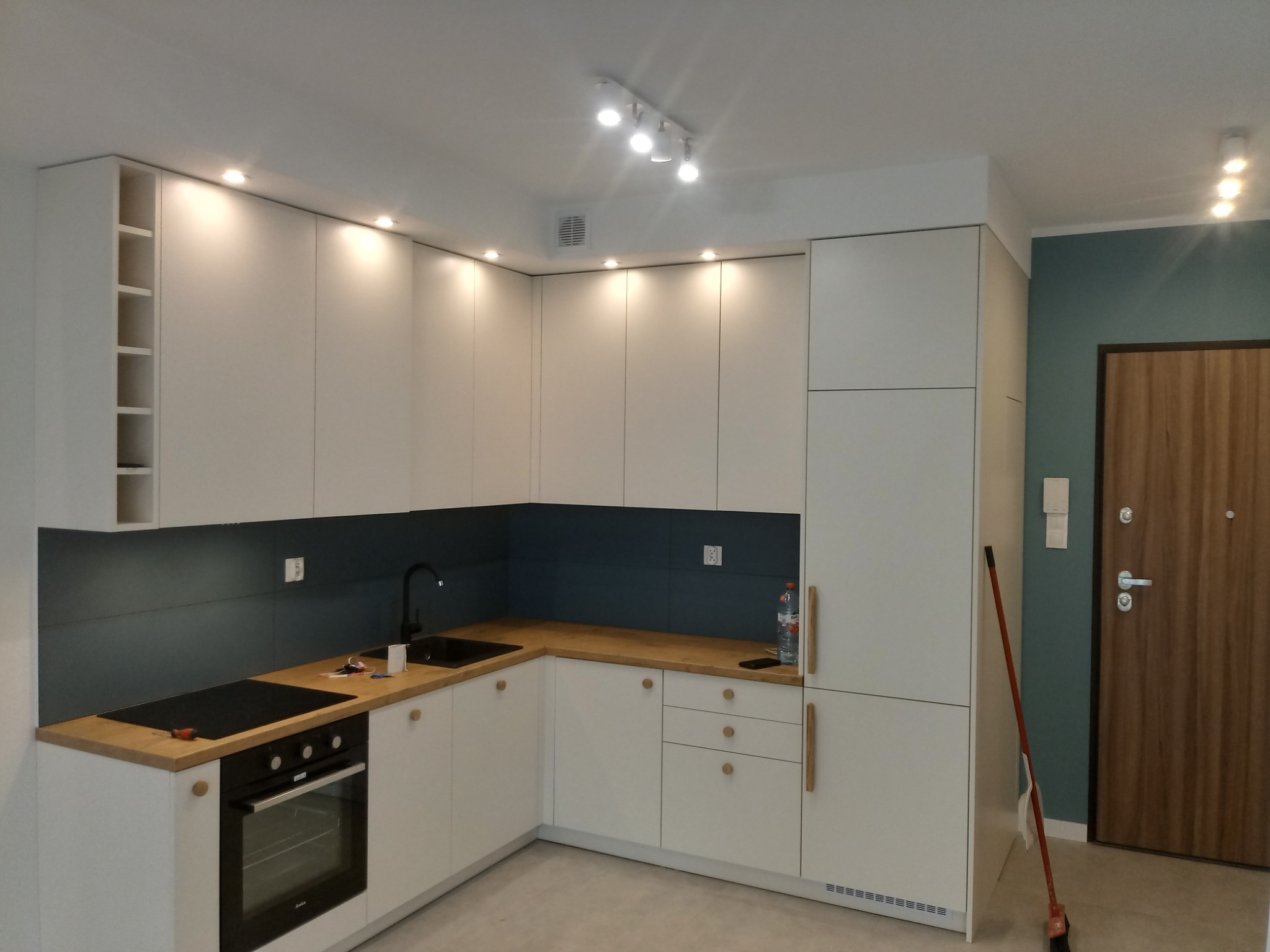 Nowoczesna kuchnia w odcieniach bieli i drewna z ciemnoniebieskim panelem nad blatem, oświetlona punktowymi lampami sufitowymi, z widocznymi narzędziami na blacie.