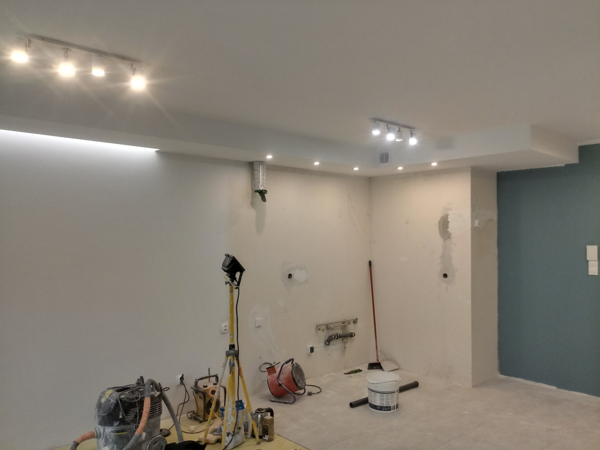 Wnętrze pokoju w trakcie remontu: ściany częściowo zagruntowane, widoczne narzędzia malarskie, odkurzacz przemysłowy, lampa na statywie, fragment pomalowanej na niebiesko ściany.