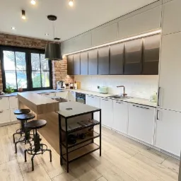 Nowoczesna kuchnia z wyspą, białymi szafkami dolnymi i górnymi z ciemnymi frontami, oświetleniem LED pod szafkami, ceglaną ścianą i drewnianą podłogą.