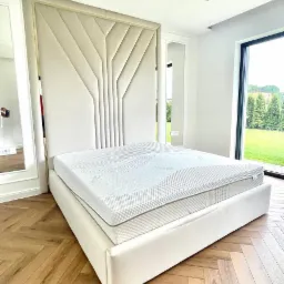 Nowoczesne łóżko z tapicerowanym zagłówkiem w geometryczne wzory, jasna kolorystyka, parkiet w jodełkę, widok na ogród przez duże okno.