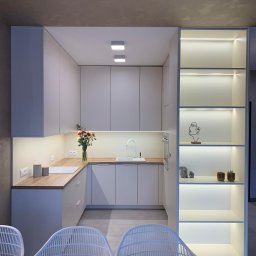 IAVIS comfort Sp.z o. o. - Nowoczesna kuchnia w odcieniach szarości z drewnianym blatem, oświetleniem LED pod szafkami i otwartym regałem z dekoracjami, widziana z perspektywy jadalni z białymi krzesłami.