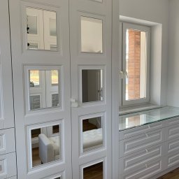 IAVIS comfort Sp.z o. o. - Biała szafa z lustrzanymi kwadratami na drzwiach, odbijającymi wnętrze garderoby z pufą; obok komoda z przeszklonym blatem i oknem w tle.