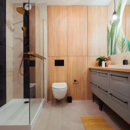 IAVIS comfort Sp.z o. o. - Nowoczesna łazienka z kabiną prysznicową typu walk-in, drewnianymi szafkami, szarą szafką pod umywalkę z czarnymi uchwytami i wiszącą toaletą.