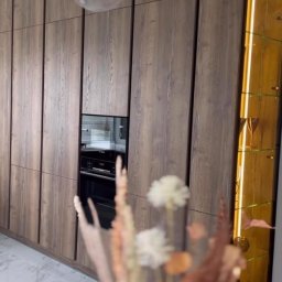 IAVIS comfort Sp.z o. o. - Nowoczesna zabudowa kuchenna z forniru w odcieniach brązu, z wbudowanym piekarnikiem i podświetlaną witryną, uzupełniona dekoracyjnym bukietem na pierwszym planie i designerską lampą wiszącą.