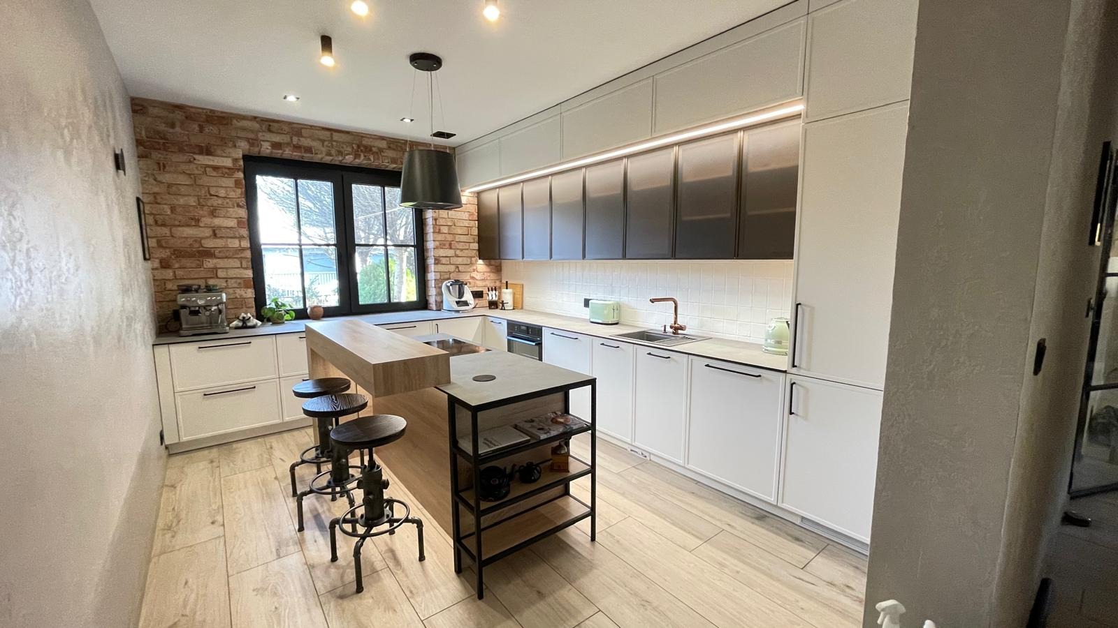Nowoczesna kuchnia z wyspą, białymi szafkami dolnymi i górnymi z ciemnymi frontami, oświetleniem LED pod szafkami, ceglaną ścianą i drewnianą podłogą.