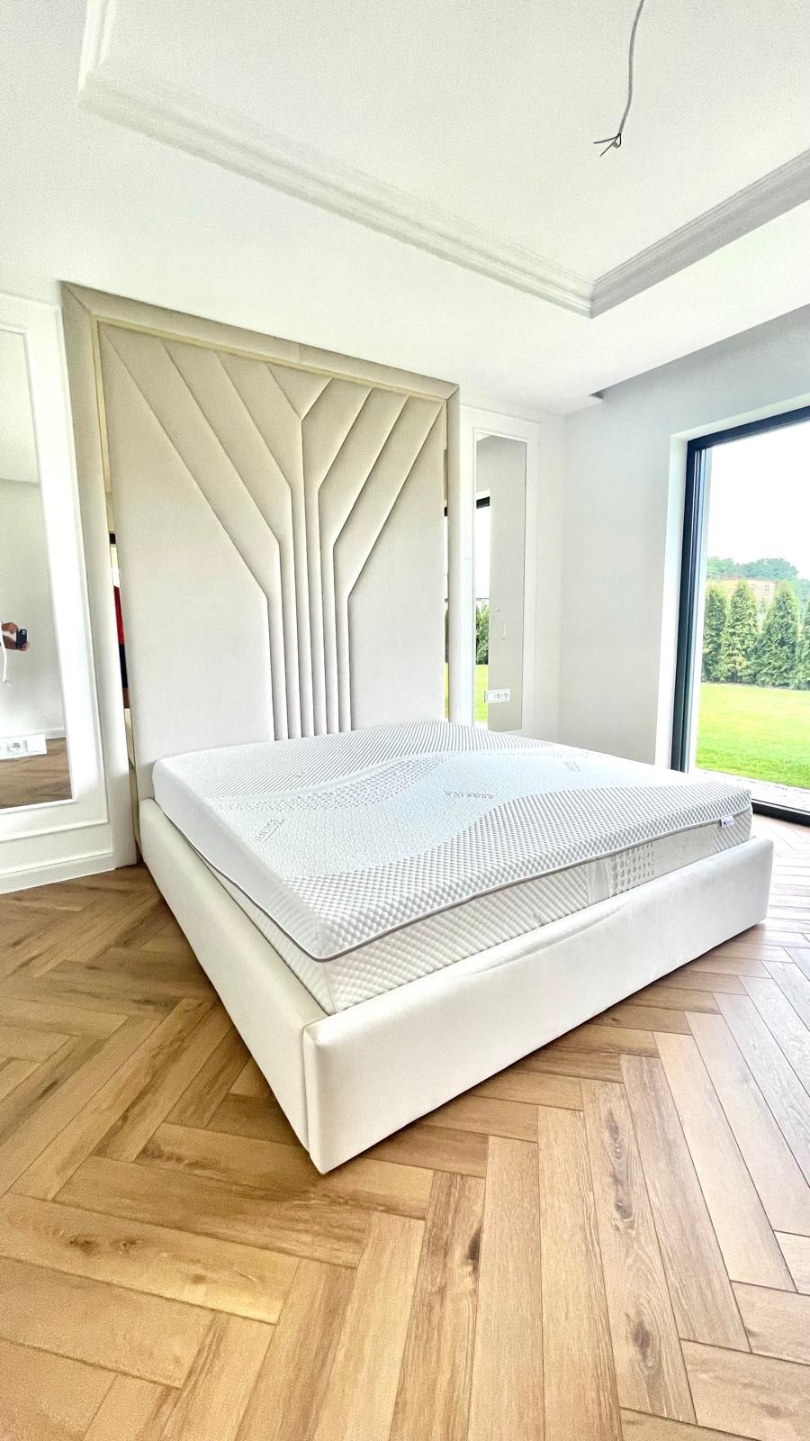 Nowoczesne łóżko z tapicerowanym zagłówkiem w geometryczne wzory, jasna kolorystyka, parkiet w jodełkę, widok na ogród przez duże okno.