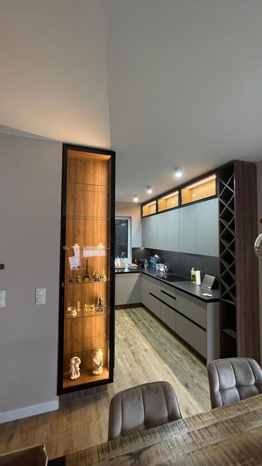 Nowoczesna kuchnia z szarymi frontami, czarnym blatem i drewnianym regałem na wino oraz przeszklona witryna z podświetleniem i dekoracyjnymi figurkami.
