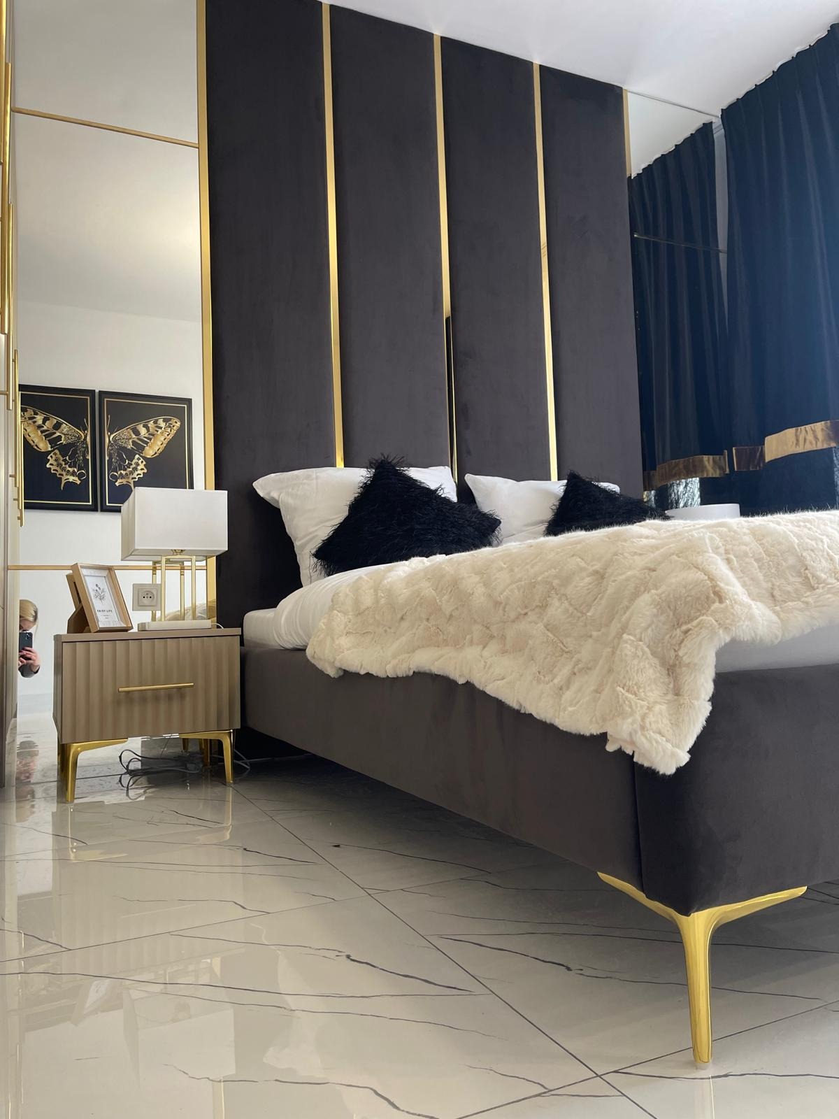 Nowoczesna sypialnia z ciemnoszarym panelem tapicerowanym za łóżkiem, akcentowanym złotymi listwami; obok stolik nocny z lampką i ramką na zdjęcia, odbicie w lustrze.