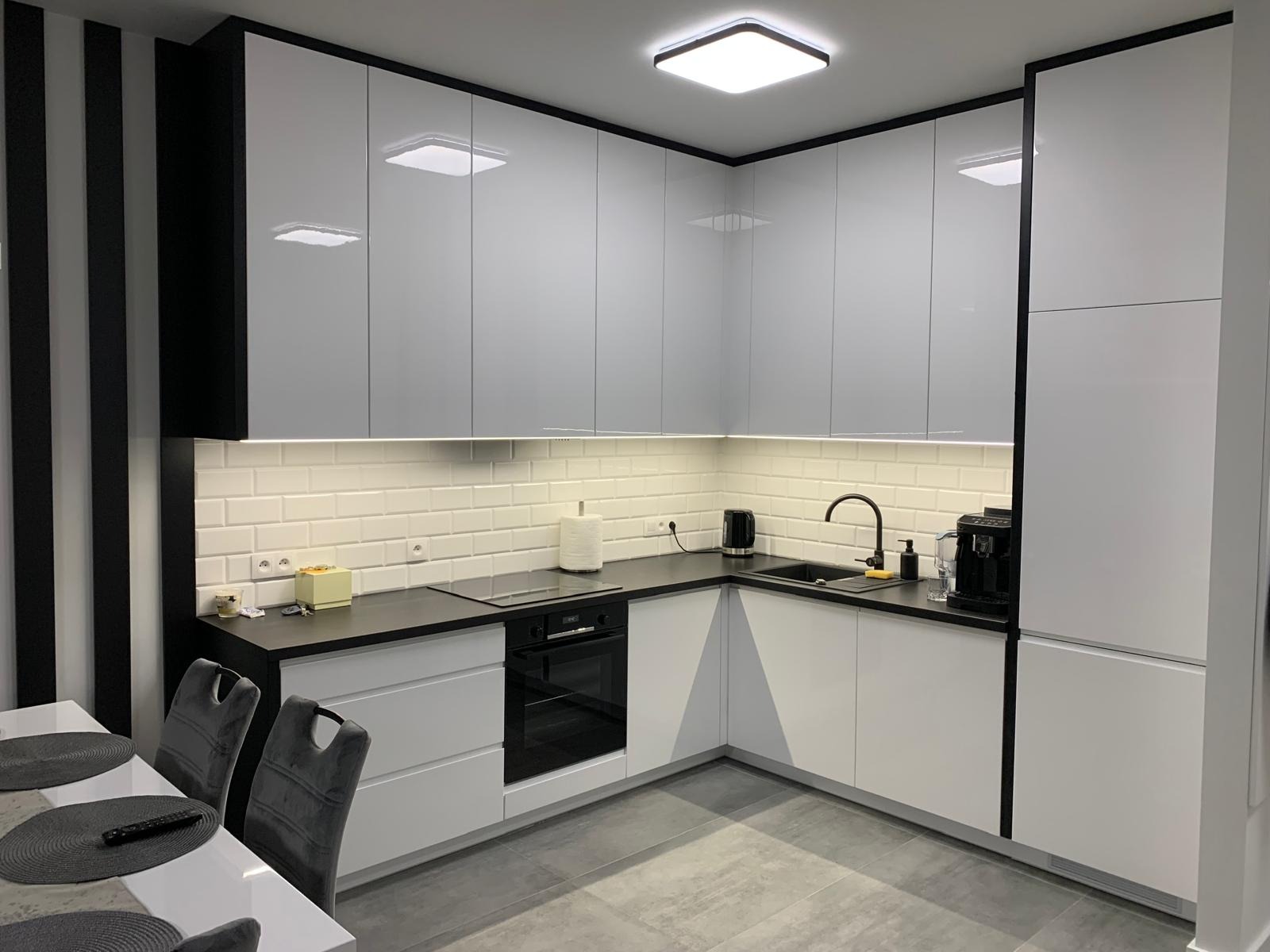 Nowoczesna kuchnia z białymi, lakierowanymi frontami szafek, czarnym blatem i elementami dekoracyjnymi w paski. Widoczny sprzęt AGD: płyta indukcyjna, piekarnik, ekspres do kawy i czajnik.