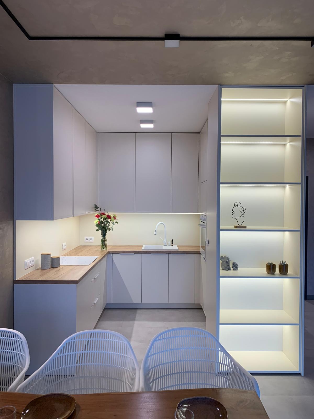 Nowoczesna kuchnia w odcieniach szarości z drewnianym blatem, oświetleniem LED pod szafkami i otwartym regałem z dekoracjami, widziana z perspektywy jadalni z białymi krzesłami.