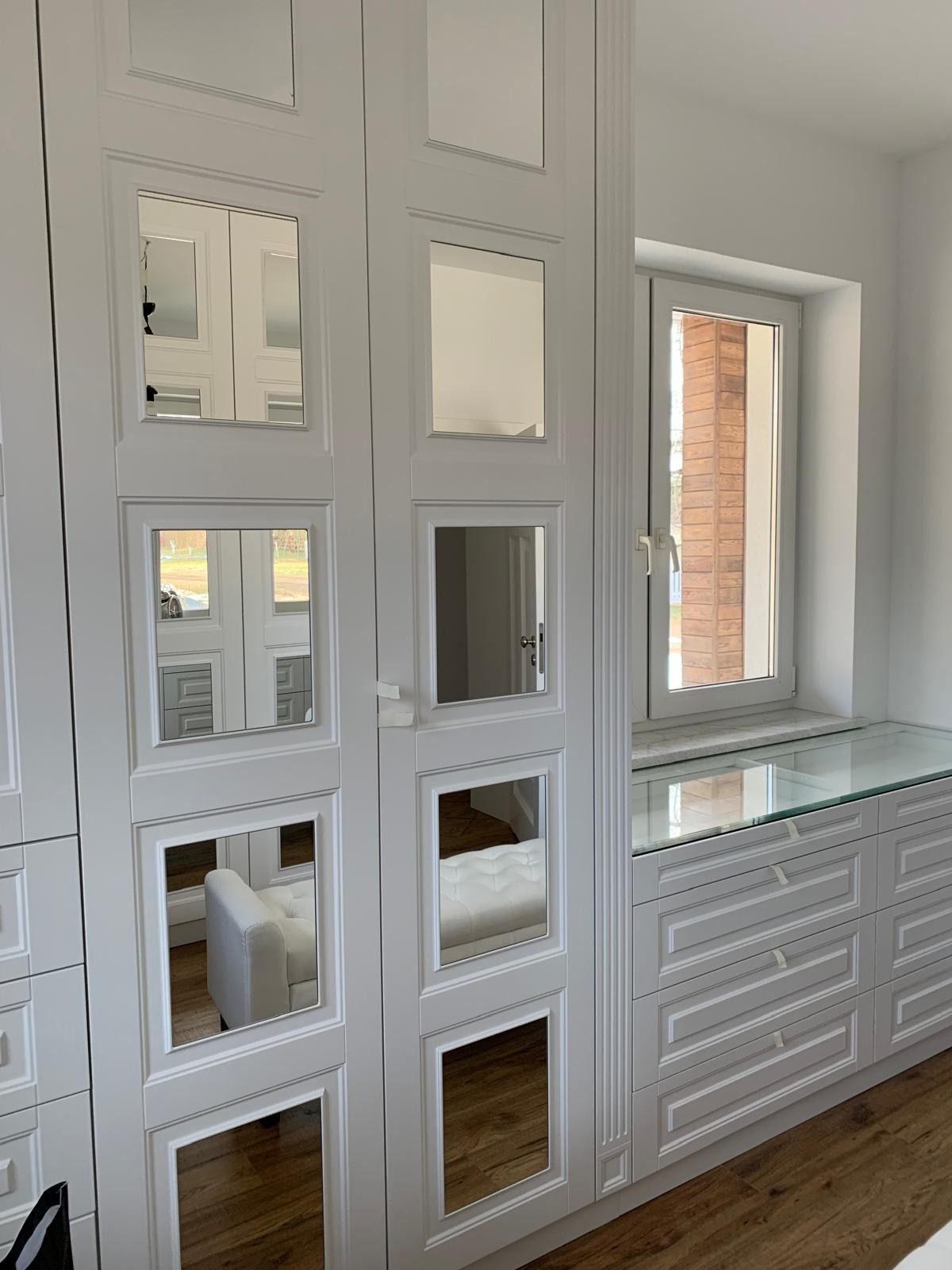 Biała szafa z lustrzanymi kwadratami na drzwiach, odbijającymi wnętrze garderoby z pufą; obok komoda z przeszklonym blatem i oknem w tle.