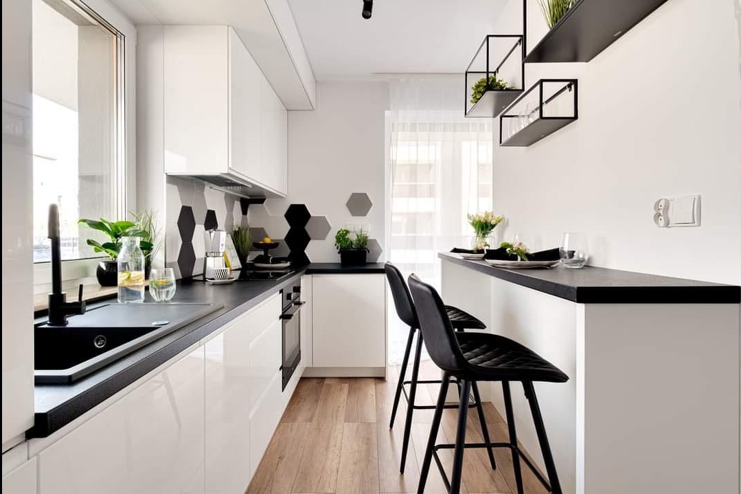 Nowoczesna kuchnia z białymi szafkami, czarnym blatem i heksagonalnymi płytkami na ścianie, uzupełniona o drewnianą podłogę i czarne hokery przy półwyspie.