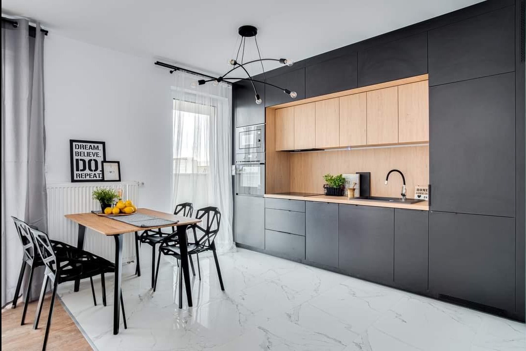 Nowoczesna kuchnia z czarnymi szafkami i drewnianymi frontami górnymi, uzupełniona blatem z czarnym zlewem i minimalistyczną armaturą, oświetlona designerską lampą wiszącą nad stołem z krzesłami...