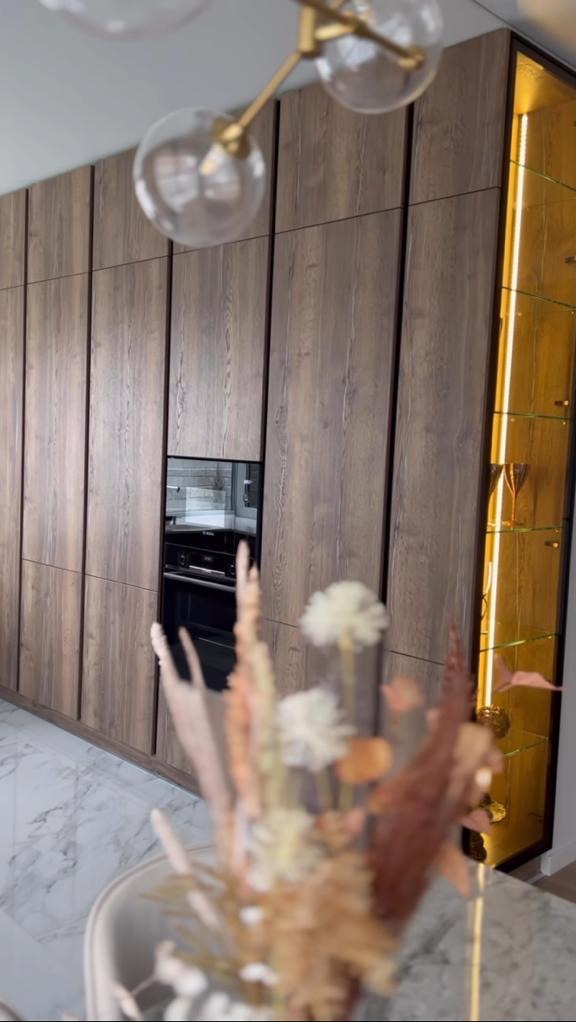 Nowoczesna zabudowa kuchenna z forniru w odcieniach brązu, z wbudowanym piekarnikiem i podświetlaną witryną, uzupełniona dekoracyjnym bukietem na pierwszym planie i designerską lampą wiszącą.