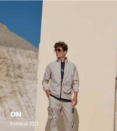 Mężczyzna w beżowym komplecie dresowym z dużymi kieszeniami cargo, pozuje na tle pustynnego krajobrazu i beżowej ściany.