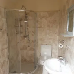 Wykończona łazienka z kabiną prysznicową, umywalką i toaletą, wyłożona płytkami w odcieniach beżu i brązu, z lustrem z małą półką.