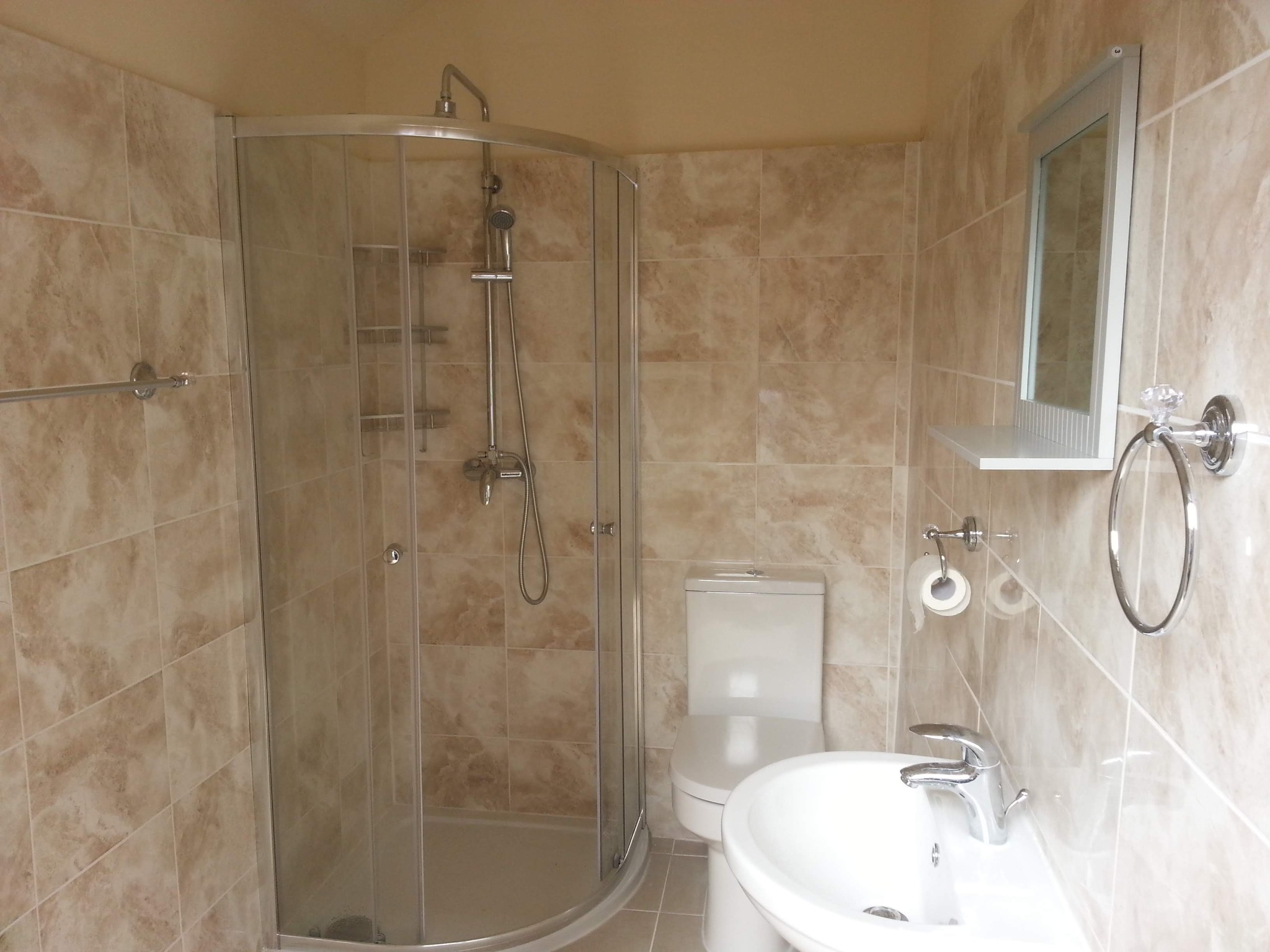 Wykończona łazienka z kabiną prysznicową, umywalką i toaletą, wyłożona płytkami w odcieniach beżu i brązu, z lustrem z małą półką.