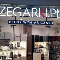 Podświetlane logo 'ZEGARKI.PL' z czerwoną literą 'K' umieszczone nad wejściem do sklepu z zegarkami, widoczne przez szklaną witrynę z ekspozycją zegarków różnych marek.