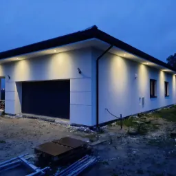 Nowoczesny dom z oświetloną elewacją i garażem, widoczne elementy rusztowania i materiały budowlane na niezagospodarowanym terenie.