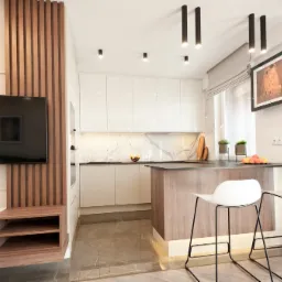 Nowoczesna kuchnia z białymi, błyszczącymi frontami szafek, marmurowym backsplashiem i wyspą z drewnopodobnym wykończeniem, oświetlona lampami sufitowymi. W tle widoczny salon z telewizorem...