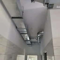 System kanałów wentylacyjnych z blachy ocynkowanej, poprowadzony pod sufitem w korytarzu o jasnych ścianach i marmurowej podłodze. Widoczne łączenia segmentów kanałów oraz elementy mocujące do sufitu.