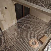 Rozłożona instalacja ogrzewania podłogowego w surowym pomieszczeniu, widoczne rury grzewcze ułożone w pętle na izolacji, okno w tle, karton i zwój rury na pierwszym planie.