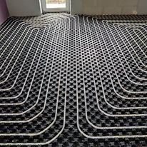 Widok z góry na ułożone rury ogrzewania podłogowego na czarnej, kwadratowej macie izolacyjnej w trakcie montażu, widoczne okno w tle.