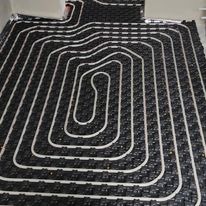 Rozłożone rury ogrzewania podłogowego na czarnej macie izolacyjnej, widok z góry, układ spiralny z meandrami, przygotowanie do wylewki.