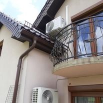 Dwa klimatyzatory zamontowane na elewacji budynku w Psarach, widoczne rynny i balkon z kutą balustradą. Jeden klimatyzator umieszczony na dachu, drugi poniżej, na ścianie.