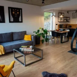 Jasne wnętrze z drewnianą podłogą, szarą sofą z żółtymi akcentami, czarnym stolikiem kawowym, wiszącym fotelem-kokonem przy kominku i otwartą kuchnią w tle.