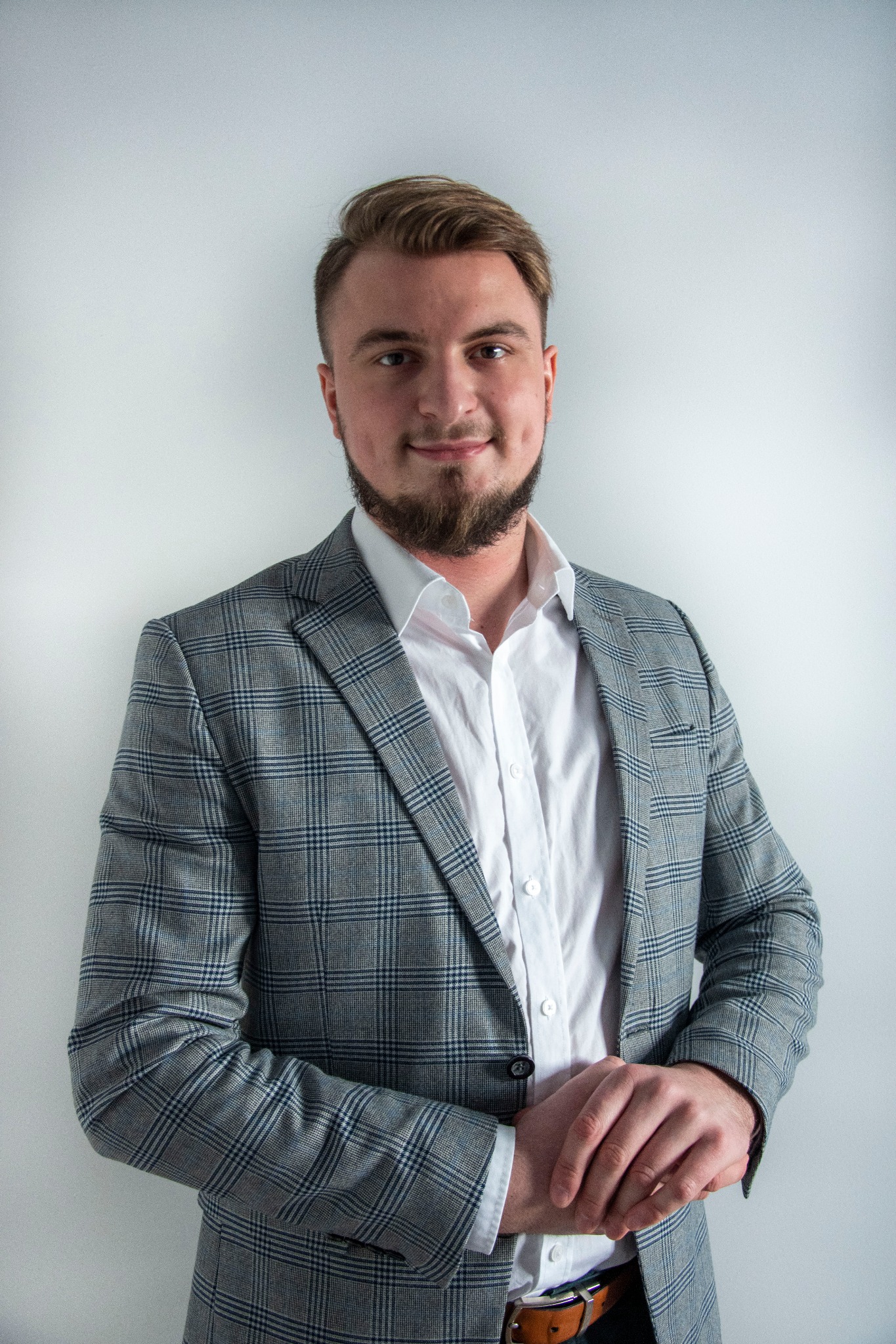 Portret mężczyzny w szarej marynarce w kratę i białej koszuli, z rękami splecionymi na wysokości pasa, na jasnym tle.