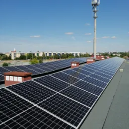 Rząd paneli słonecznych na płaskim dachu budynku z kominami wentylacyjnymi, w tle wieża telekomunikacyjna i zabudowa miejska pod błękitnym niebem.