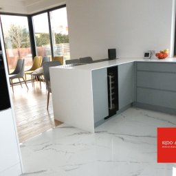 KĘDO Kędzior Dariusz - Nowoczesna kuchnia z szarymi szafkami, białą wyspą i podłogą z marmuru, wbudowana chłodziarka na wino, widok na ogród przez duże okna.